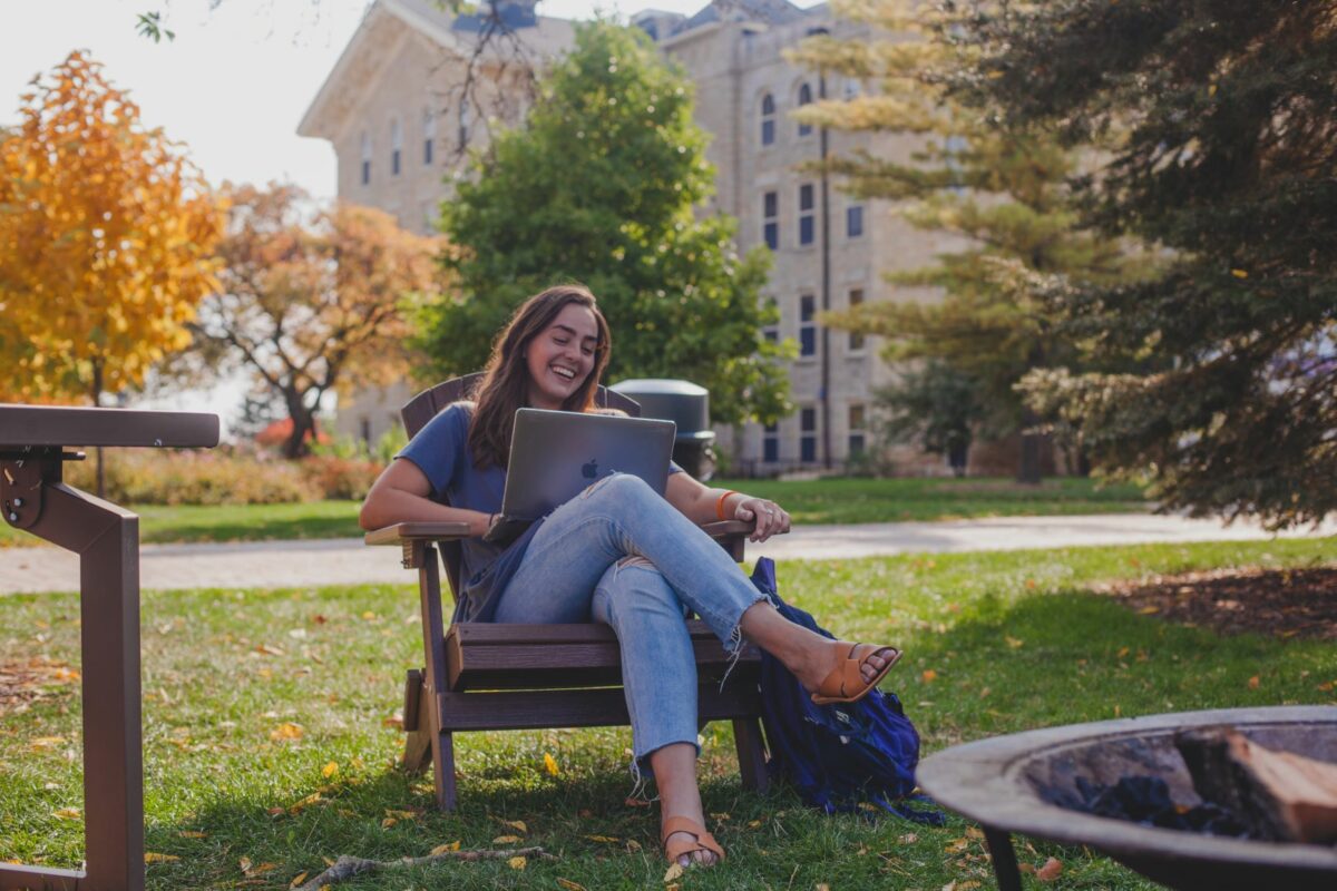 A photo of a Wheaton student outside Blanchard Hall conducting a Writing Consultation from her laptop