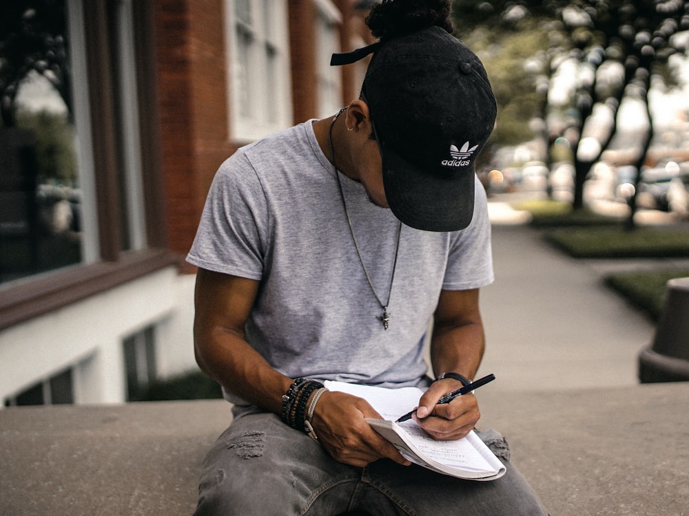 A photo of a man in a black baseball cap sitting down writing in a journal